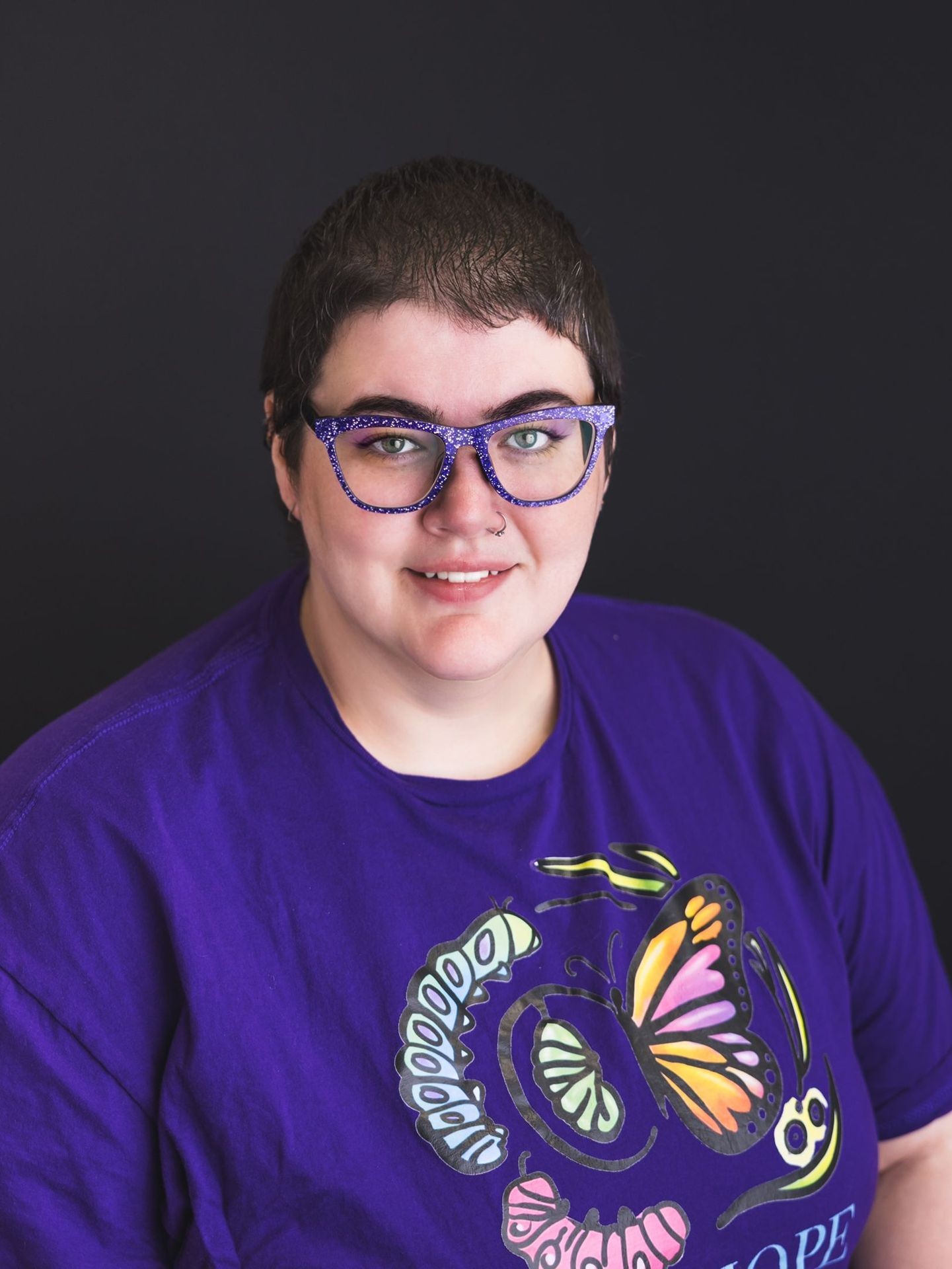 Person with short hair wearing purple glasses and a purple shirt with a colorful butterfly design.
