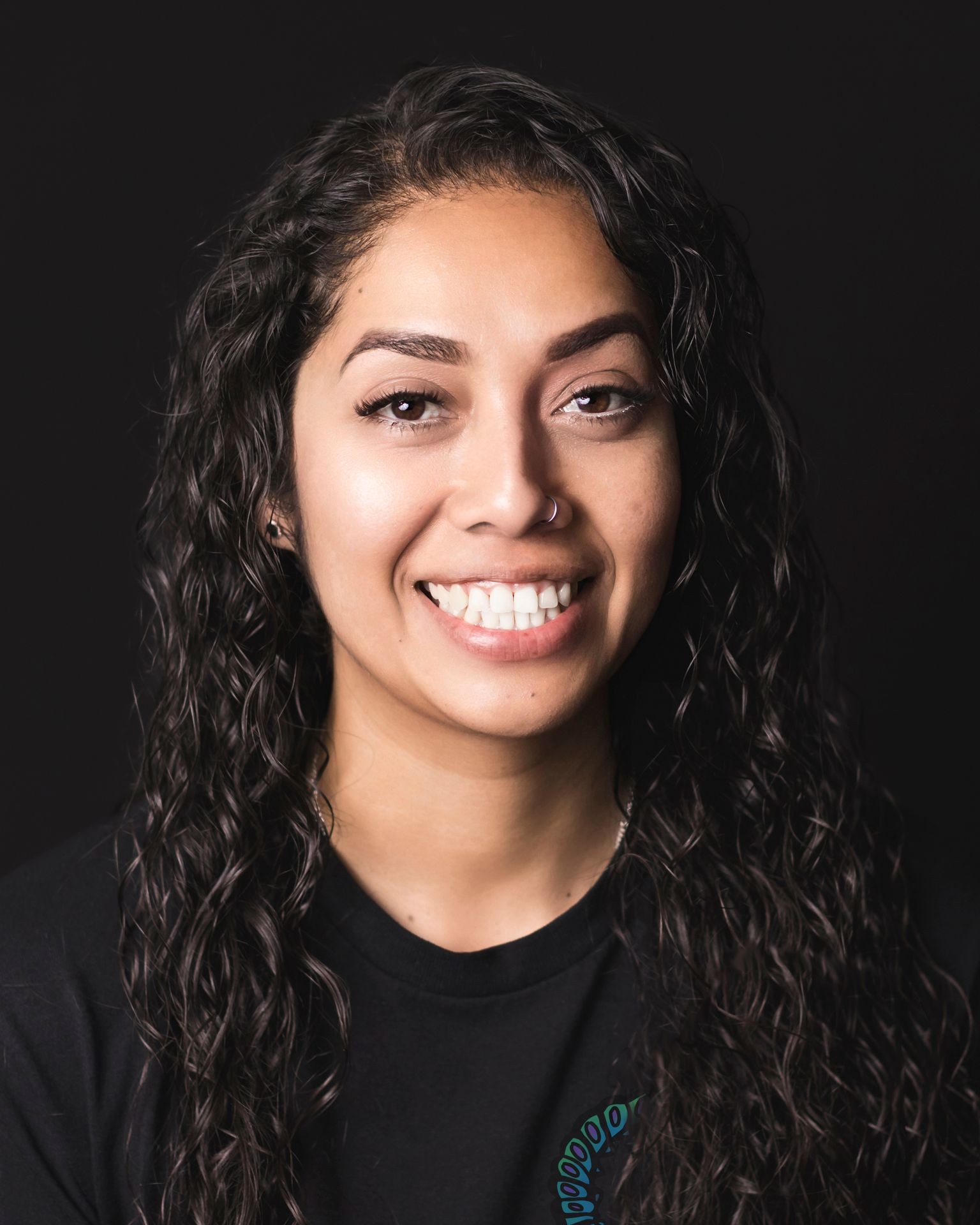 Smiling woman with wavy hair, wearing a black shirt, against a dark background.