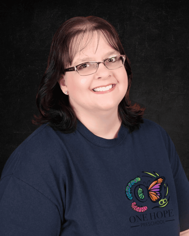 Smiling individual with glasses, wearing a navy blue One Hope Preschool shirt against a dark background.