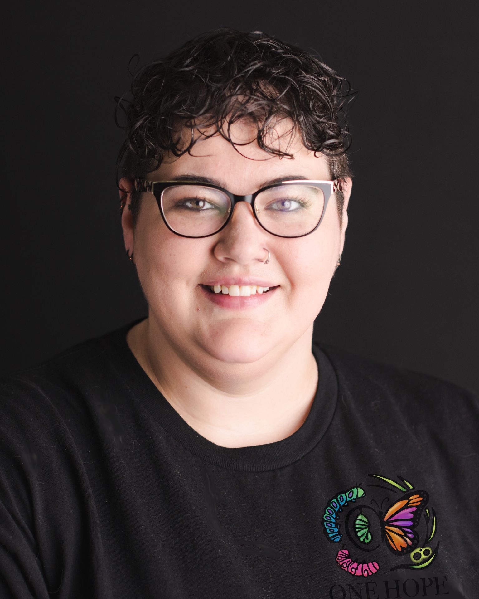 Person with short curly hair and glasses smiling, wearing a black shirt with a colorful butterfly logo.