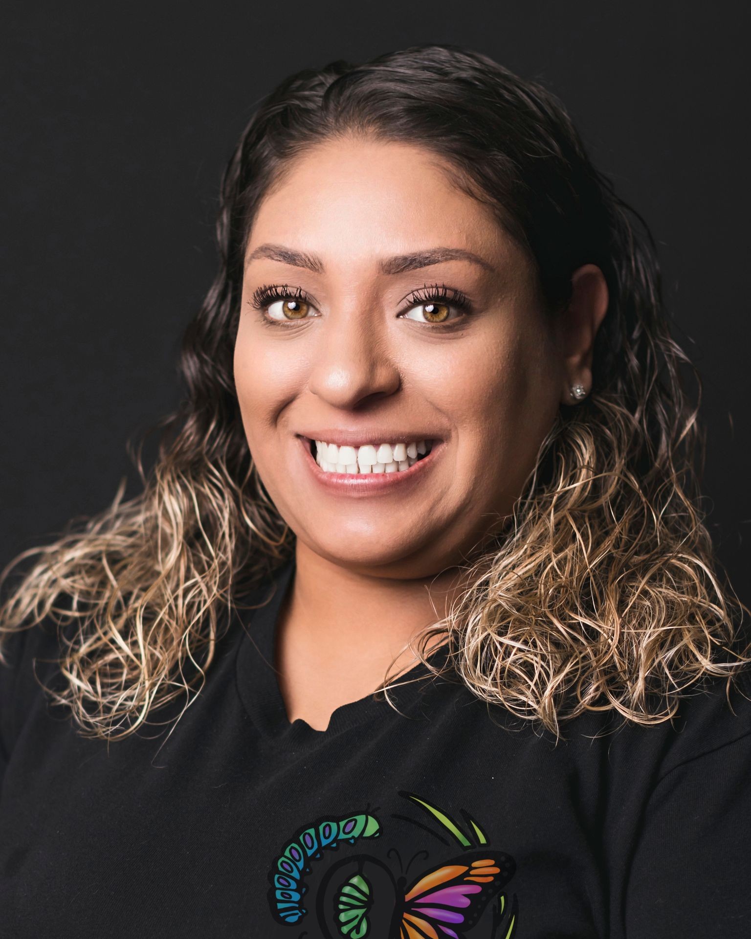 Person with wavy hair wearing a black shirt featuring a colorful butterfly design on a dark background.