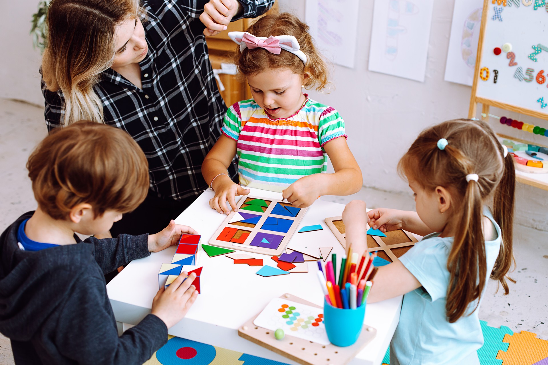 Portrait of young pretty woman sitting at table with pupils children playing. Teacher touching headband of little girl.